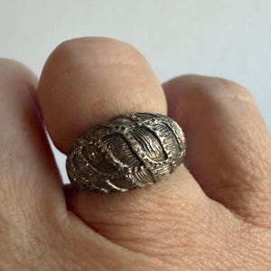 Pode incluir: Um anel prateado com um design texturizado e com nervuras. O anel tem formato oval e parece ser feito de metal. O padrão intrincado confere ao anel uma estética vintage ou antiga. O anel é usado em um dedo.