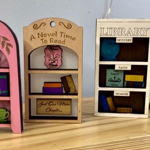 May include: Four miniature wooden bookshelves in pink, natural, and white tones. The shelves are decorated with books, a teacup, and the words "A Novel Time To Read", "Library", and "Never Ending Shelf".