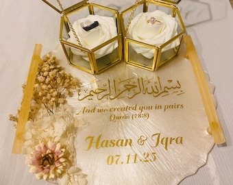 May include: Two gold-framed, hexagonal ring boxes with the letters "I" and "H" on top. Each box holds a white rose and a ring. The boxes sit on a decorative, off-white tray with gold accents and text that reads "Hasan & Iqra 07.11.25".