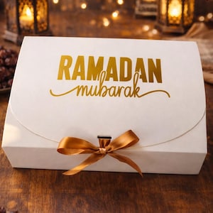 May include: A white gift box with a gold ribbon and the words "RAMADAN mubarak" in gold script. The box is on a wooden surface, with blurred lanterns and lights in the background. Dates are visible in the foreground.