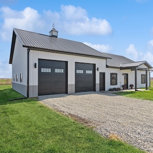 May include: A white and gray metal building with two black garage doors and a covered porch. The building has a black roof and a white trim. The building is surrounded by green grass and a gravel driveway.