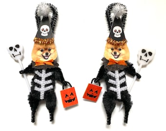 May include: Two black and white skeleton dog costumes with orange pumpkin buckets and a white skull on a stick. The dogs are wearing black and orange party hats with a skull design.
