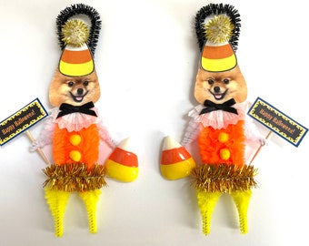 May include: Two Halloween decorations featuring a Pomeranian dog dressed as candy corn. Each figure has a candy corn hat, orange body, and holds a sign that says "Happy Halloween!". They also have a candy corn in their hand.