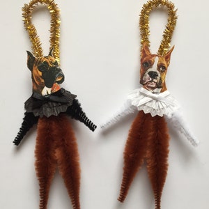 May include: Two brown pipe cleaner ornaments with boxer dog faces. One dog is wearing a black ruffled collar and the other is wearing a white ruffled collar.