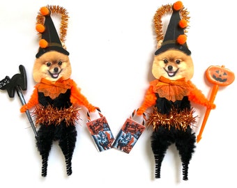 May include: Two handmade Halloween ornaments featuring a Pomeranian dog dressed in a witch costume. The ornaments are made with pipe cleaners and have orange and black accents. One ornament holds a black cat and a bag of candy, while the other holds a pumpkin on a stick.