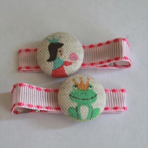 May include: Two pink hair clips with fabric buttons. One button features a princess with a crown and a pink flower. The other button features a green frog with a gold crown.