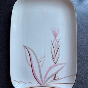 Op de afbeelding: Rechthoekige witte serveerschaal met een bloemmotief in roze, bruin en grijs. De schaal heeft afgeronde hoeken en een bruine rand. Het bloemmotief toont gestileerde bladeren en een centrale bloem.