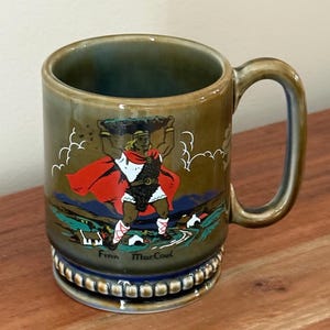 Puede incluir: Una taza de cerámica con asa, con una colorida ilustración de una figura con capa roja que lleva una cesta. La taza tiene un esmalte verde y marrón con el texto "Finn MacCool" en la base. La taza está sobre una superficie de madera.