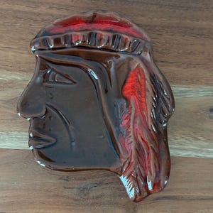Puede incluir: Una pieza decorativa de cerámica marrón con forma de perfil nativo americano. El tocado y el cabello presentan detalles en rojo. La pieza es brillante y descansa sobre una superficie de madera.