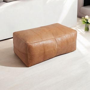 May include: A rectangular brown leather ottoman with a stitched seam down the center.