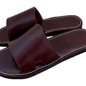 May include: A pair of brown leather sandals with a white stitch detail around the edges. The sandals have a simple design and are made of high-quality leather.