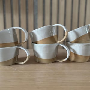 Peut inclure: Six tasses en céramique blanche et brune avec des anses. Les tasses sont empilées par paires, la tasse du haut de chaque paire ayant un glaçage blanc et la tasse du bas un glaçage brun.