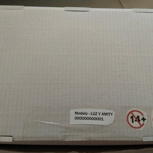 K&ouml;nnte beinhalten: Rechteckiger Karton in hellem Beige mit ausgeschnittenen Laschen oben. Ein Aufkleber zeigt den Text "Modelo - LUZ Y AMITY" und eine Seriennummer. Zwei Warnsymbole sind ebenfalls auf der Box zu sehen.