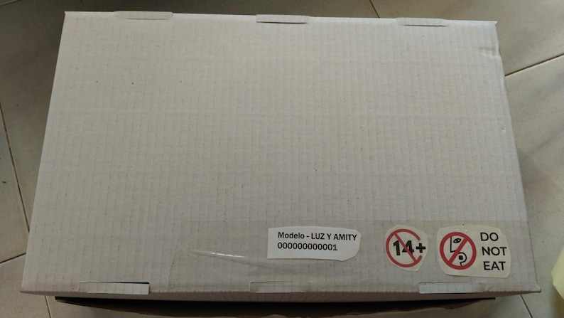 K&ouml;nnte beinhalten: Rechteckiger Karton in hellem Beige mit ausgeschnittenen Laschen oben. Ein Aufkleber zeigt den Text "Modelo - LUZ Y AMITY" und eine Seriennummer. Zwei Warnsymbole sind ebenfalls auf der Box zu sehen.