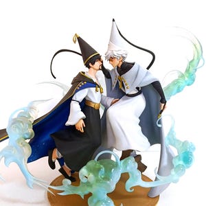 Puede incluir: Una figura detallada que muestra a dos personajes con sombreros puntiagudos y capas fluidas, uno en blanco y negro y el otro en blanco y azul claro. Están rodeados por efectos de energía azul claro translúcidos y arremolinados sobre una base circular.