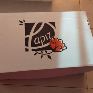K&ouml;nnte beinhalten: Wei&szlig;er Karton mit einem Logo mit dem Wort "apiz" in stilisierter Schrift, begleitet von einem floralen Design in Orange und Schwarz. Die Box ist rechteckig und geschlossen.