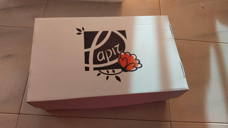 K&ouml;nnte beinhalten: Wei&szlig;er Karton mit einem Logo mit dem Wort "apiz" in stilisierter Schrift, begleitet von einem floralen Design in Orange und Schwarz. Die Box ist rechteckig und geschlossen.