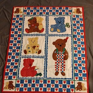 May include: A rectangular quilt featuring a blue and white chequered border with red hearts. The centre displays four panels with teddy bears: two brown and tan bears, a blue patterned bear, a yellow bear with patches, and a brown bear in overalls.