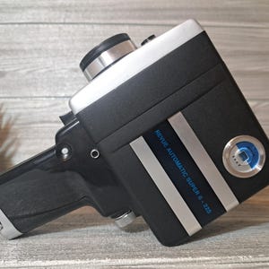 Può includere: Una cinepresa vintage Revue Automatic Super 8-225. La fotocamera è nera con dettagli argentati e bianchi. Il testo "Revue Automatic Super 8-225" è stampato sul lato. Ha un'impugnatura a pistola e un obiettivo.