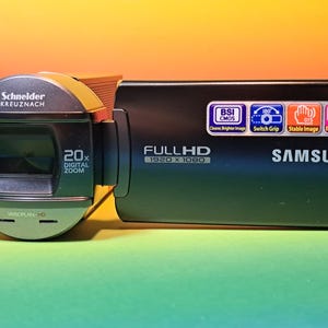 Pode incluir: Câmara de vídeo Samsung preta com lente Schneider Kreuznach. Possui zoom digital de 20x e grava em Full HD 1920 x 1080. A câmara de vídeo tem ícones de função, incluindo "BSI CMOS" e "Estabilização de imagem".