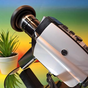 May include: A silver and black vintage video camera with a black lens cover. The camera has a black strap and is sitting on a reflective surface.