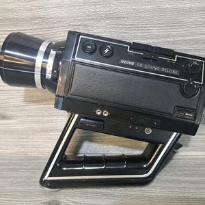 Può includere: Telecamera cinematografica vintage Revue 8 Sound Deluxe nera, con un obiettivo grande e una maniglia. La fotocamera ha un corpo nero con dettagli argentati e diversi quadranti e pulsanti. Il testo "Revue 8 Sound Deluxe" è stampato sopra.