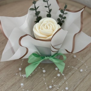 Puede incluir: Una vela blanca con forma de rosa se presenta en papel blanco con bordes marrones. Ramitas verdes y una cinta verde claro añaden color. Un collar de perlas decora la superficie de madera, creando un delicado arreglo floral.