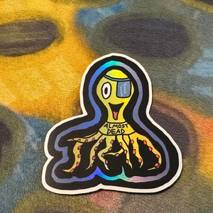 May include: A yellow octopus with one eye and a band around its head that says "Almost Dead". The octopus is drawn in a cartoon style and has a black outline. The background is a blurry image of a colorful fabric.