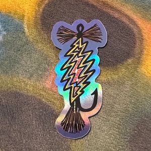 May include: A holographic sticker of a lightning bolt shaped fishing lure with red, white, and blue accents. The lure has a black hook and a brown and black tassel.