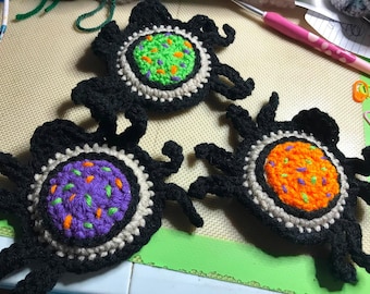 May include: Three handmade crochet spider coasters featuring black yarn legs and a central circular body. The centers are textured in bright green, purple, and orange with contrasting speckles, each bordered by a ring of light beige yarn.