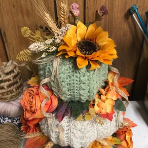 May include: A stacked decorative pumpkin arrangement featuring three crocheted pumpkins in sage green and cream colours. The pumpkins are adorned with artificial sunflowers, orange roses, wheat stalks, and autumn leaves, accented with thin twine ribbons.