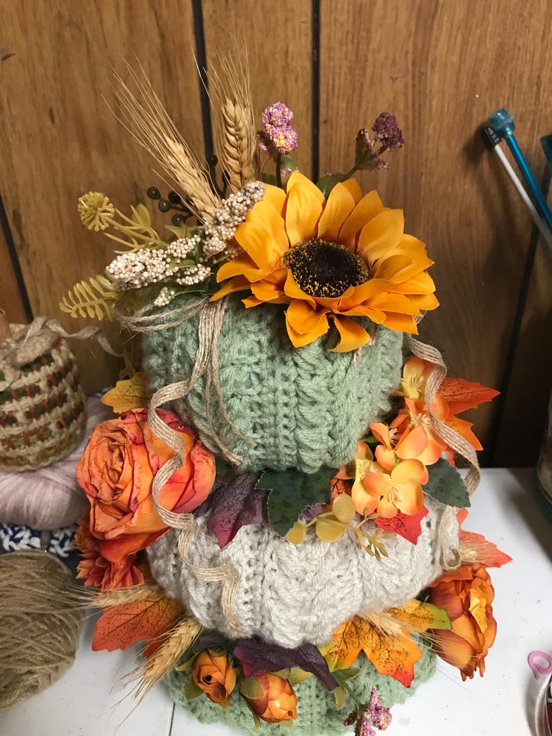 May include: A stacked decorative pumpkin arrangement featuring three crocheted pumpkins in sage green and cream colours. The pumpkins are adorned with artificial sunflowers, orange roses, wheat stalks, and autumn leaves, accented with thin twine ribbons.