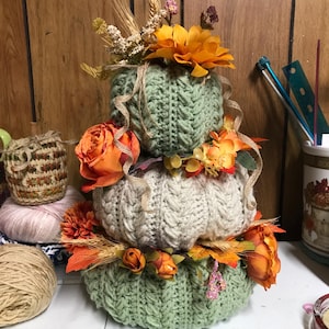 May include: A stacked trio of crocheted pumpkins in sage green and cream, featuring a cable knit texture. The pumpkins are decorated with artificial orange flowers, dried wheat stalks, and thin brown ribbons tucked between each layer.