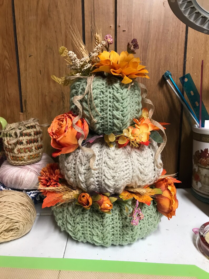 May include: A stacked trio of crocheted pumpkins in sage green and cream, featuring a cable knit texture. The pumpkins are decorated with artificial orange flowers, dried wheat stalks, and thin brown ribbons tucked between each layer.