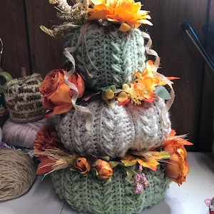 May include: A stacked trio of crocheted pumpkins in sage green and cream, featuring a cable knit texture. The pumpkins are decorated with artificial orange roses, yellow flowers, dried wheat stalks, and thin burlap ribbons wrapped around them.