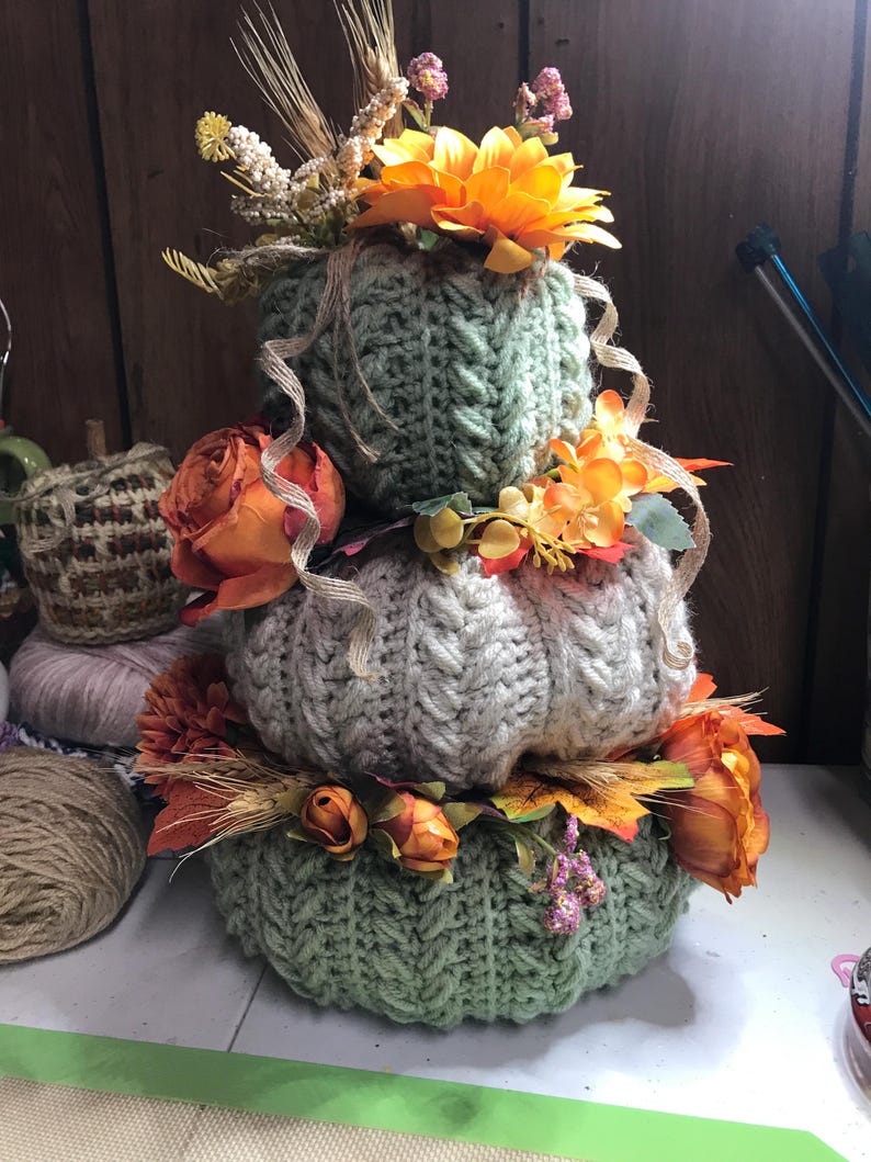 May include: A stacked trio of crocheted pumpkins in sage green and cream, featuring a cable knit texture. The pumpkins are decorated with artificial orange roses, yellow flowers, dried wheat stalks, and thin burlap ribbons wrapped around them.