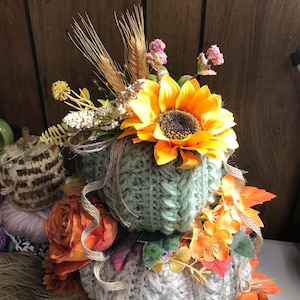 May include: A stacked decorative pumpkin set featuring two crocheted pumpkins in sage green and cream. The pumpkins are adorned with artificial sunflowers, orange roses, wheat stalks, and small pink flowers, accented with thin strands of jute twine.