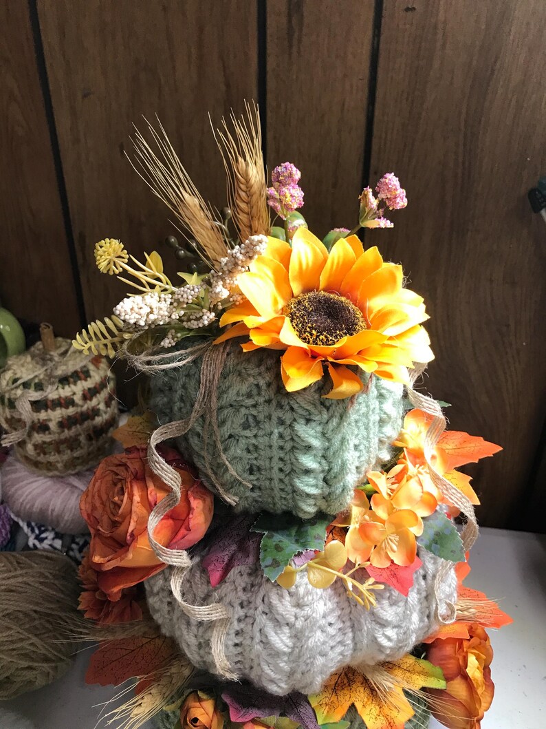 May include: A stacked decorative pumpkin set featuring two crocheted pumpkins in sage green and cream. The pumpkins are adorned with artificial sunflowers, orange roses, wheat stalks, and small pink flowers, accented with thin strands of jute twine.