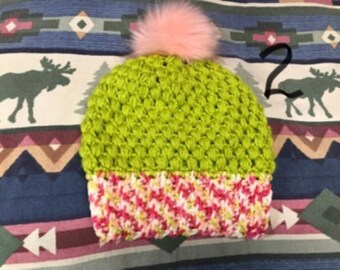 May include: A lime green crocheted beanie with a textured bobble stitch pattern, featuring a wide cuff made of variegated pink, white, and yellow yarn. The top of the hat is finished with a fluffy, light pink pom-pom. The number 2 is visible.
