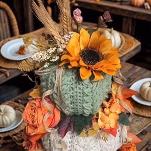 Könnte beinhalten: Eine gestapelte Kürbisdekoration bestehend aus einem salbeigrünen Strickkürbis, der mit einer großen orangefarbenen Sonnenblume, Weizenähren und kleinen weißen Blüten verziert ist, auf einem cremefarbenen Strickkürbis mit orangefarbenen Stoffrosen.