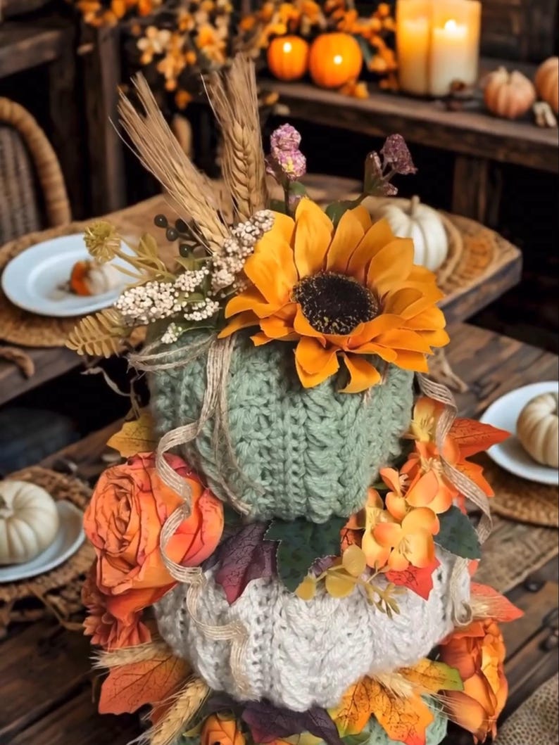 May include: A stacked pumpkin decoration featuring a sage green knitted pumpkin topped with a large orange sunflower, wheat stalks, and small white flowers, resting on a cream-coloured knitted pumpkin adorned with orange fabric roses and autumn leaves.