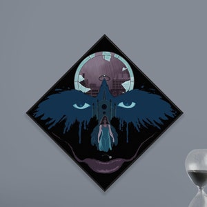 May include: A framed artwork with a dark, stylized illustration of a face with blue wings and eyes, a church, and a figure in a dress. The artwork is displayed on a gray wall above a black shelf with books, a pink sphere, and an hourglass.