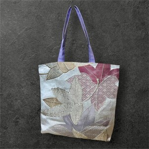 May include: A white tote bag with a floral design featuring purple, pink, and gold leaves. The bag has lavender straps.