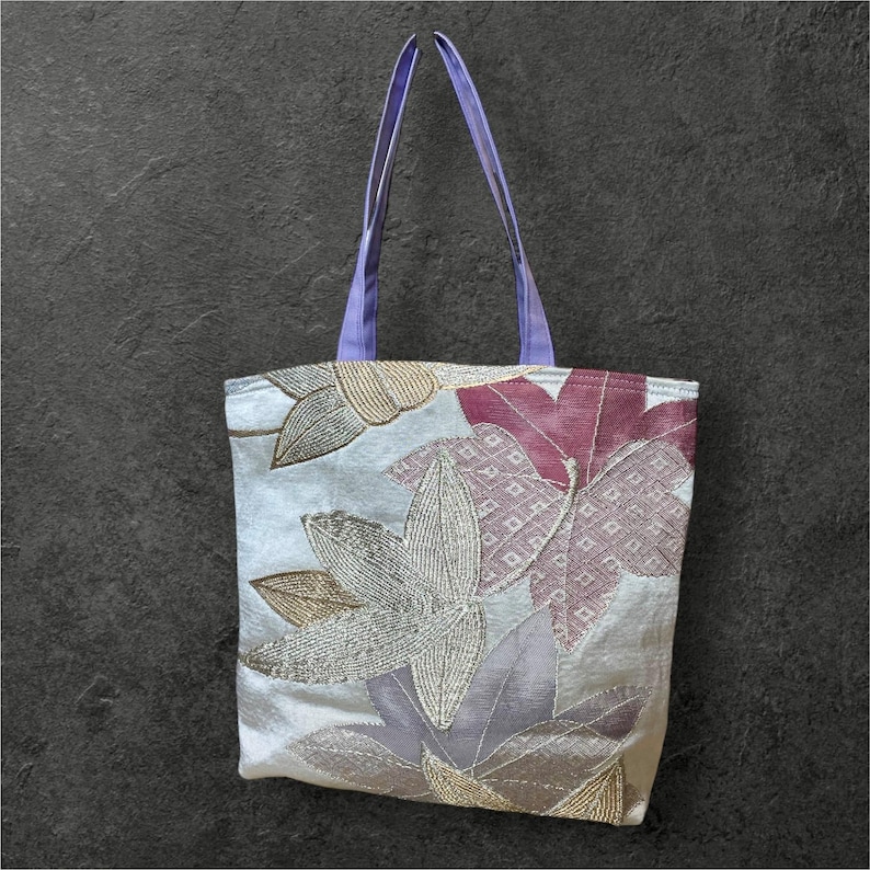 May include: A white tote bag with a floral design featuring purple, pink, and gold leaves. The bag has lavender straps.