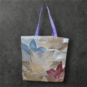 May include: A white tote bag with a floral design featuring blue, gold, and red leaves. The bag has lavender straps.