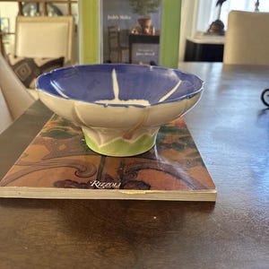 May include: A ceramic bowl with a scalloped rim, featuring a deep blue interior that transitions into a white exterior with a green base. The bowl has a distinct floral-inspired shape with white petal-like accents extending from the center.
