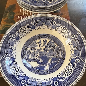 May include: Two round ceramic bowls featuring a blue and white willow pattern. The design includes a central landscape with a bridge, pagoda, trees, and birds, surrounded by a wide, intricate geometric and floral border on the rim of each bowl.