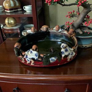 May include: A decorative ceramic bowl with a dark green interior and a red base. The bowl is adorned with small figurines of people in various poses around the rim. A bonsai tree with red berries is in the background.