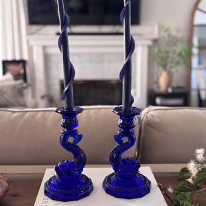 May include: Two cobalt blue glass candle holders with a dolphin design, each holding a dark purple spiral candle. The candle holders are placed on a white book titled "Jacques Cousteau The Ocean World".