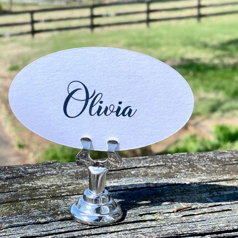 May include: A white oval place card with the name "Olivia" in elegant cursive script, held by a shiny silver metal holder. The holder has a decorative design and a round base. The background shows a wooden surface and a blurred outdoor scene.